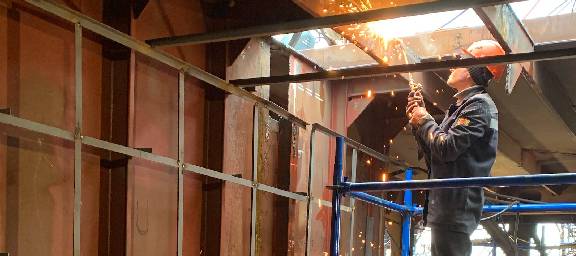 Maintenance technician performing welding repairs to a rusted metal structure with light and obvious breach overhead, while standing on a blue scaffolding, wearing an orange safety hat and blue coveralls.